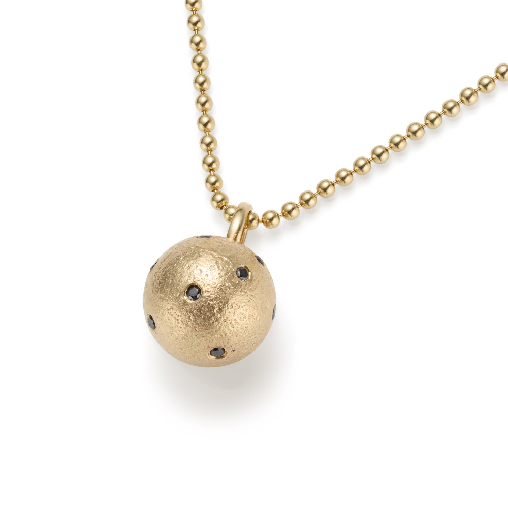 Close-up of a gold moon pendant, textured and dotted with black diamonds, hanging on a gold ball chain. The pendant's unique surface and precious stones contrast strikingly against the black background.